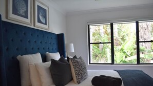 A bed with a blue headboard, white bedding, and multiple pillows. There is a window with a view of greenery outside. - Blue Belle - Walk to Beach, Golf & Cafe`s, slps 7 (Shelley Beach)