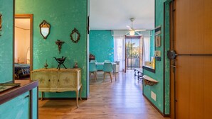 A hallway with teal walls, a wooden floor, and a door on the right. There is a table with chairs in the background, and a ceiling fan is visible. - Il Giardino by Wonderful Italy (Sanremo)