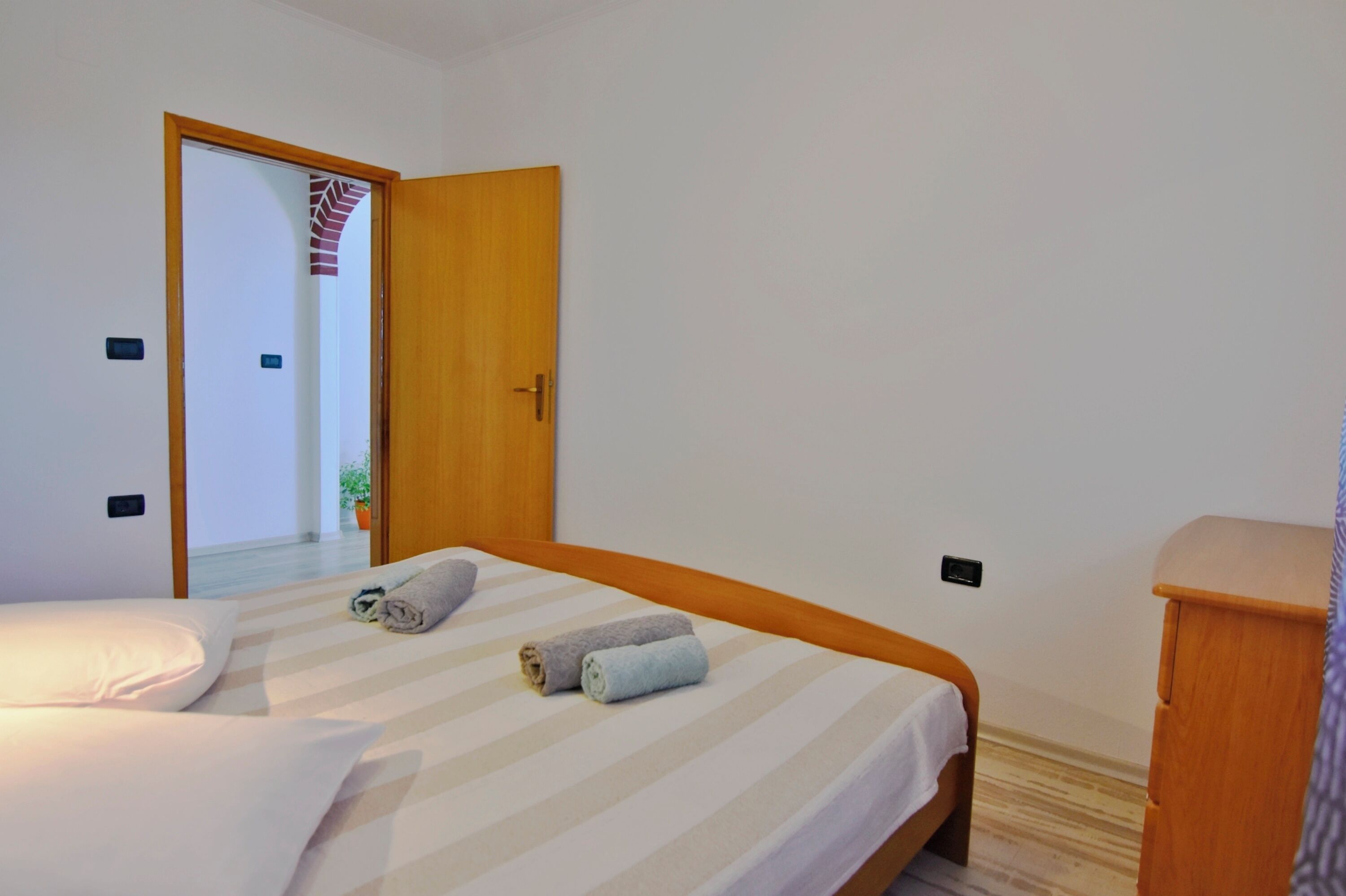 A hotel room with a bed, a wooden door, a small wooden dresser, and a potted plant in the hallway.