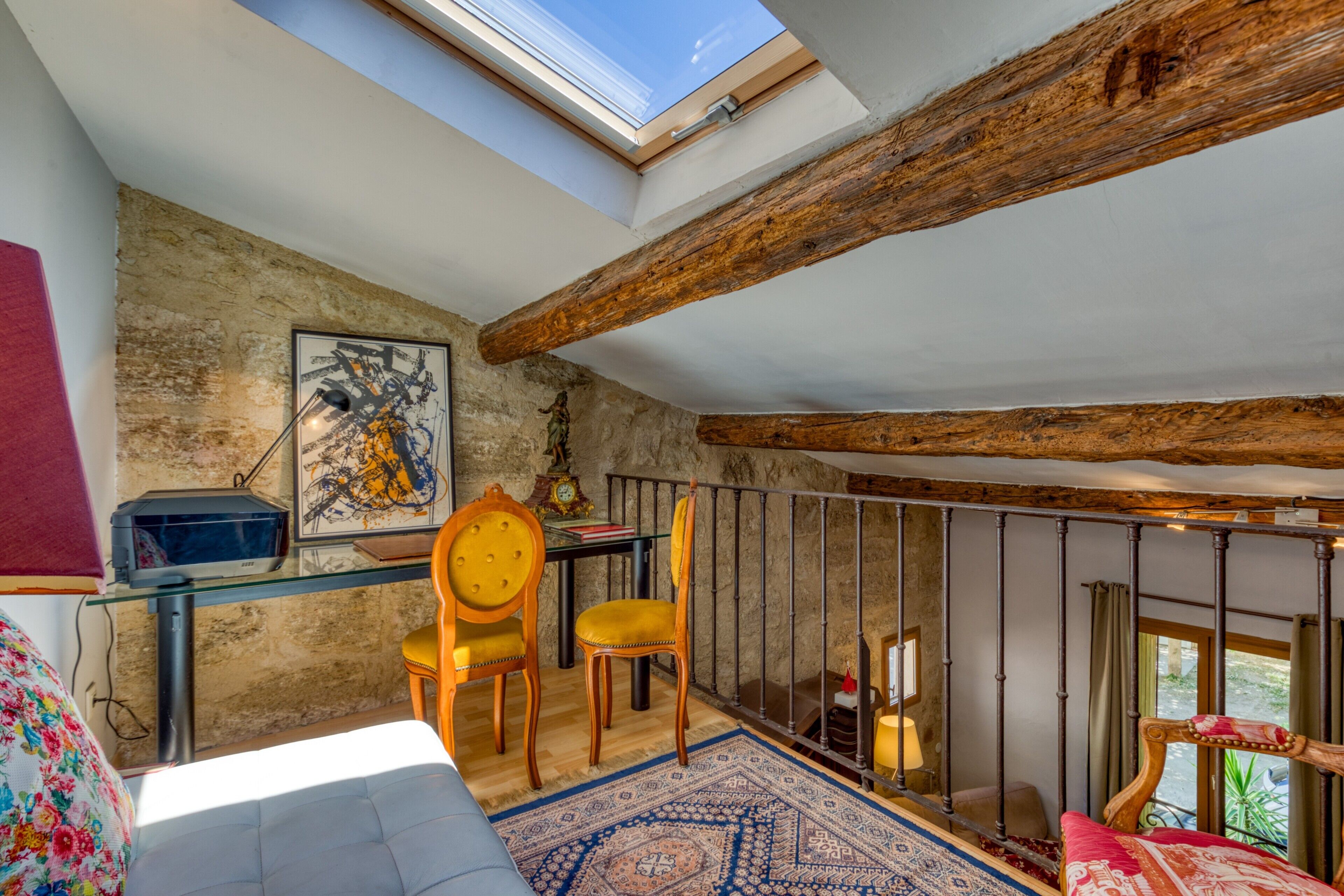 A room with a skylight, rustic wooden beams, a glass table, two chairs, a patterned rug, and a bed with a red and white bedspread.