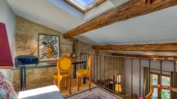 Une pièce dotée d’une lucarne, de poutres en bois rustiques, d’une table en verre, de deux chaises, d’un tapis à motifs et d’un lit recouvert d’une couvre-lit rouge et blanche.