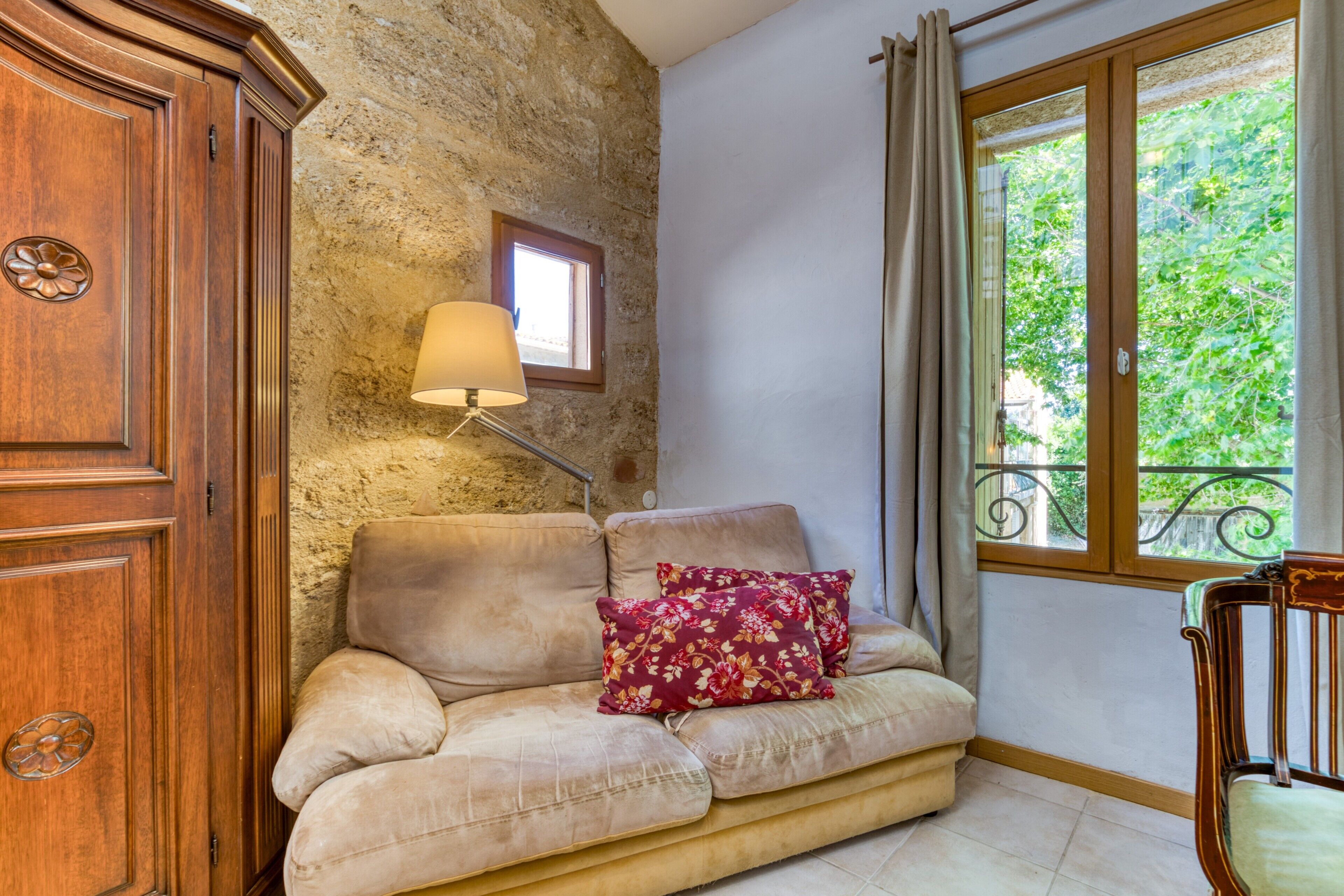 Ein Zimmer mit Steinsandsteinwand, einem Holzschrank, einer Couch mit Blumenkissen, einem Tisch und einem Fenster mit Blick ins Grüne.