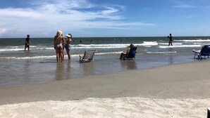 Een strand met mensen die staan en zitten op het zand, een paar stoelen en de oceaan op de achtergrond.
