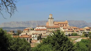 A town with a prominent church featuring a bell tower, surrounded by residential buildings with red-tiled roofs, set against a backdrop of mountains. - Provencal village between Aix en Provence and Cassis (Fuveau)