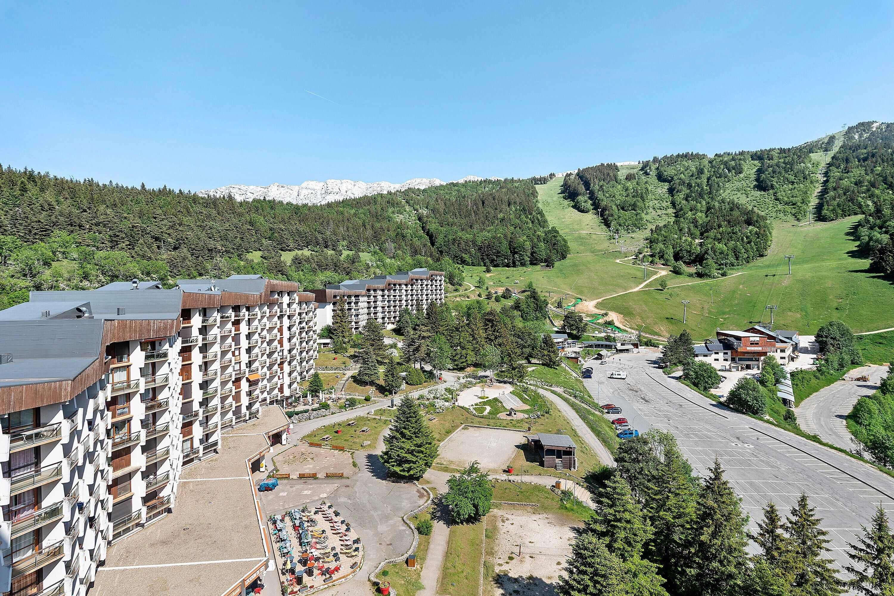 Een meerlaags hotel met balkons, een parkeerplaats en uitzicht op een bergachtig landschap.