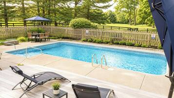 A rectangular outdoor swimming pool with a wooden deck, two lounge chairs, a small table with a plant, and a wooden fence in the background.
