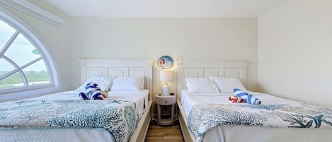 Ein Hotelzimmer mit zwei Betten, einem Deckenventilator, einem Nachttisch mit Lampe und Blick durch ein Fenster.