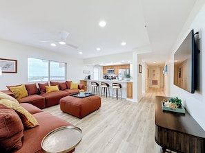 A modern living room with a sofa, ottoman, and coffee table, a kitchen with bar stools, and a large TV mounted on the wall. - Recently renovated BEACH condo, tons of amenities (Indian Shores)