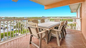 Ein Balkon mit Holztisch und -stühlen, Blick auf eine Straße und Grünflächen, und einem strahlend blauen Himmel.
