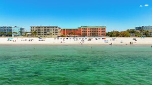 Ein Hotel direkt am Strand mit mehreren Etagen, ein sandiger Strand im Vordergrund und ein klarer, blauer Himmel.