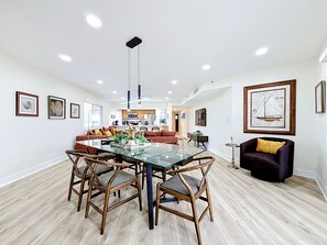Ein moderner Essbereich mit Glastisch und Holzkohlen, ein Wohnzimmer mit Sofa und Kissen sowie eine Küche im Hintergrund.