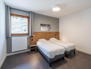 Quarto com duas camas, janela com cortinas, radiador, quadro na parede e luminária no teto.
