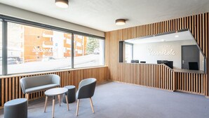 A reception area with a wooden panel wall, a reception desk with a mirror, two grey chairs, a small round table, and a large window with a view of buildings and parked cars. - 6-guest Alpine Rental With Balcony (Auris)