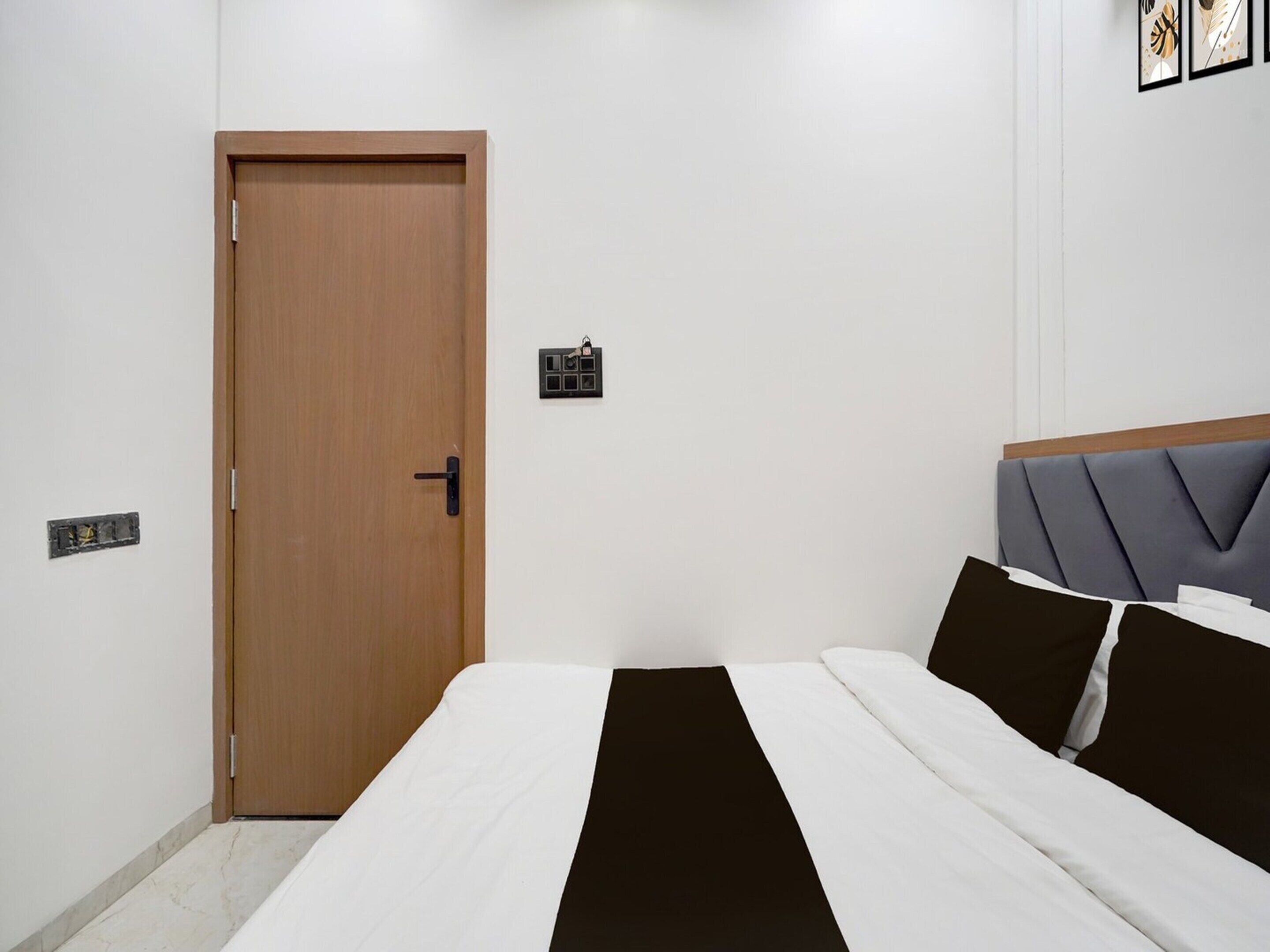 A bedroom with a wooden door, a bed with white and black bedding, a grey upholstered headboard, and a small wall-mounted light switch.