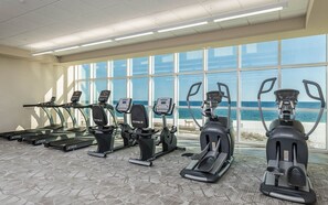 Un gimnasio con cintas de correr, elípticas y una bicicleta estática, con amplias ventanas y vista al mar.