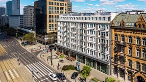 Un edificio de varios pisos con amplias ventanas, una zona peatonal con mesas al aire libre y una intersección de calles.