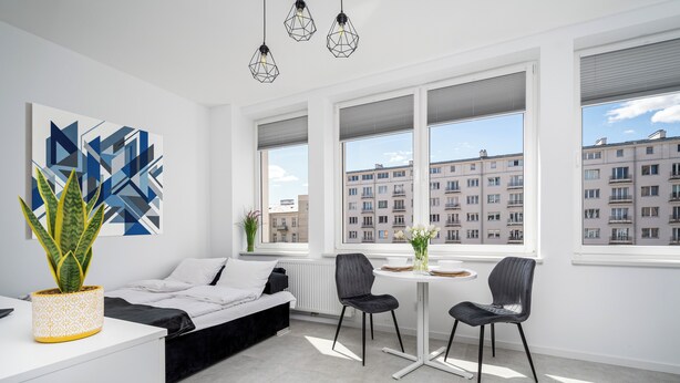 Un dormitorio moderno con cama, una mesa de comedor con sillas, una planta en maceta y ventanas amplias con vista a la ciudad.