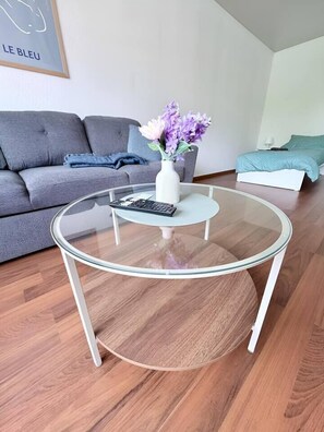 Ein modernes Wohnzimmer mit Glastisch, grauem Sofa und einem Bett mit blauer Decke.
