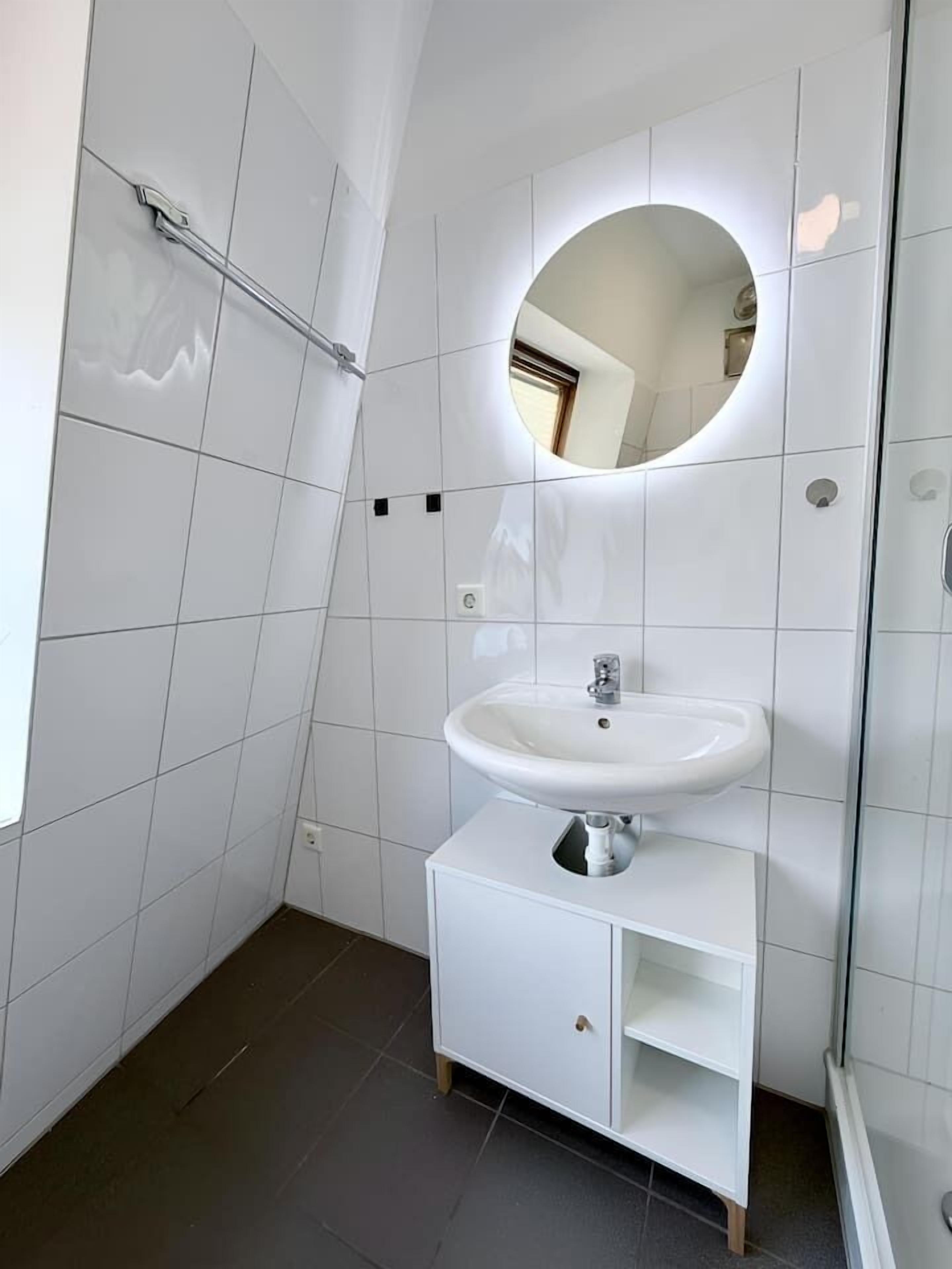 Un bany modern amb rajoles blanques, un mirall rodó, un lavabo amb pedestal i un moble amb calaixos.