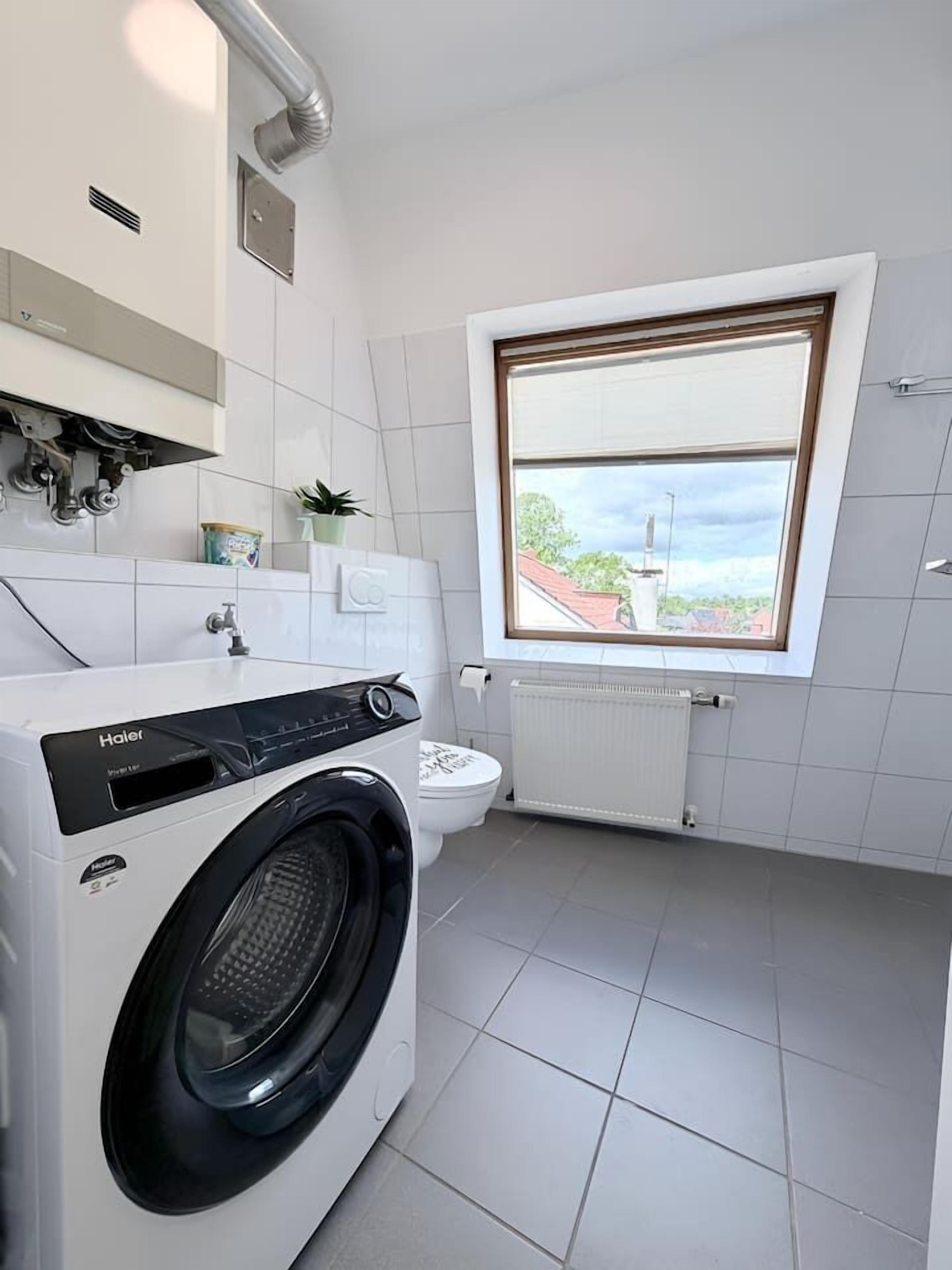 Un baño moderno y compacto con lavadora de carga frontal, inodoro, radiador y ventana con vistas al exterior.