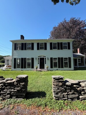 A two-story building with a central entrance, stone walls in the foreground, and a clear blue sky.