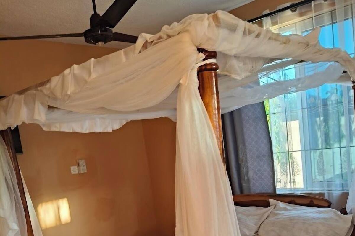 Una cama con dosel de madera, un ventilador de techo y un ventanal con cortinas.