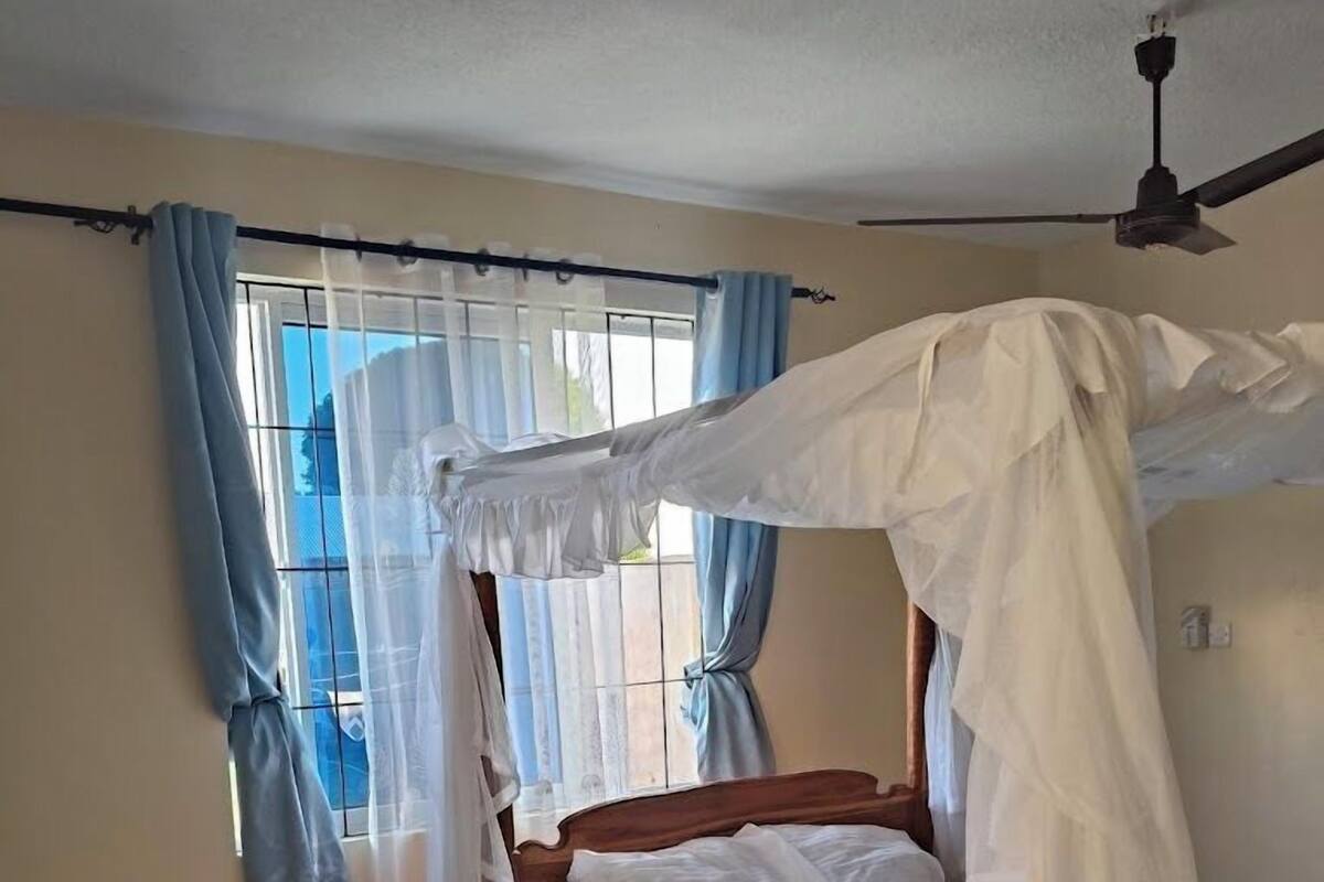 Un dormitorio con una cama de madera, un ventilador de techo, una ventana con cortinas y un tapete estampado.