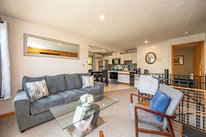 Ein Wohnzimmer mit einer Couch, einem Couchtisch und einem Sessel. Im Hintergrund ist eine Küche zu sehen.