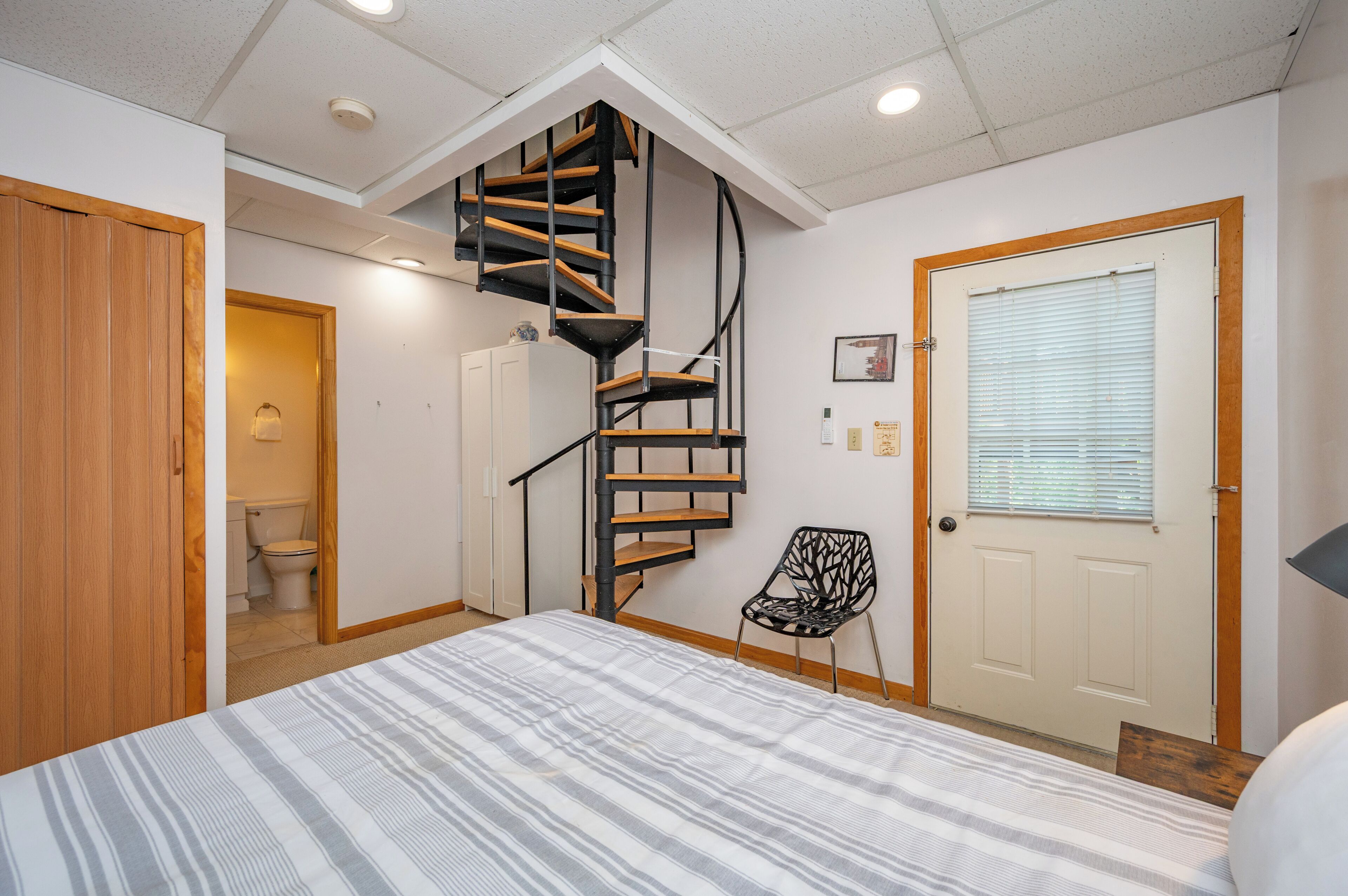 A bedroom with a spiral staircase, a bed, a chair, and a door.