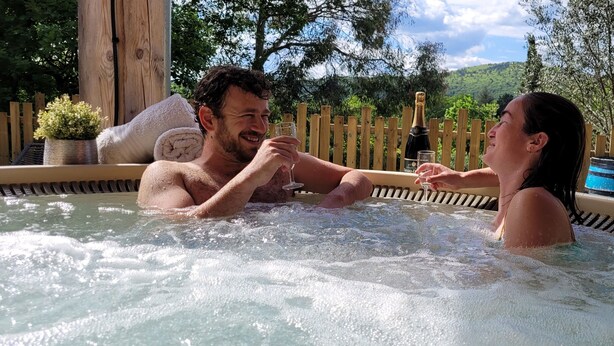 Una pareja en un jacuzzi con una botella de champán, rodeada por una valla de madera y follaje.
