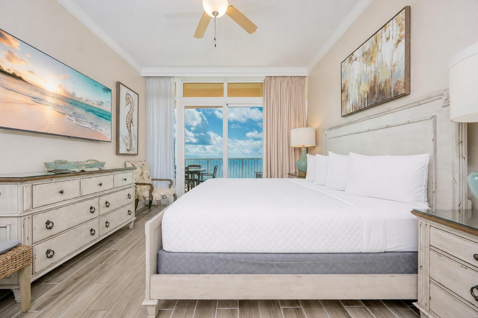 A hotel room with a large bed, a dresser, a chair, a table, and a view of the sky and ocean through the windows.