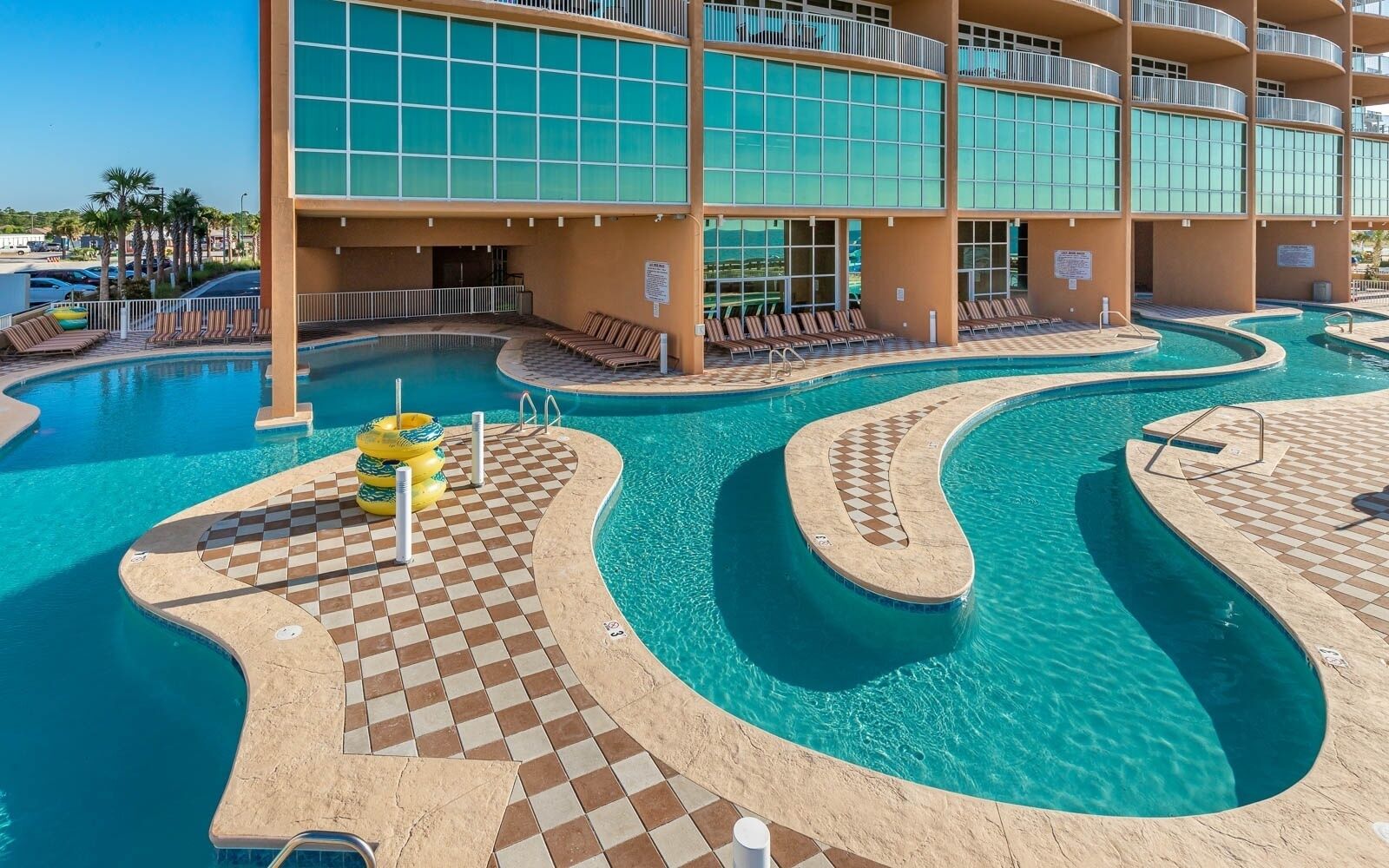A hotel pool area with multiple swimming pools, lounge chairs, and a checkered tile pattern around the pools.