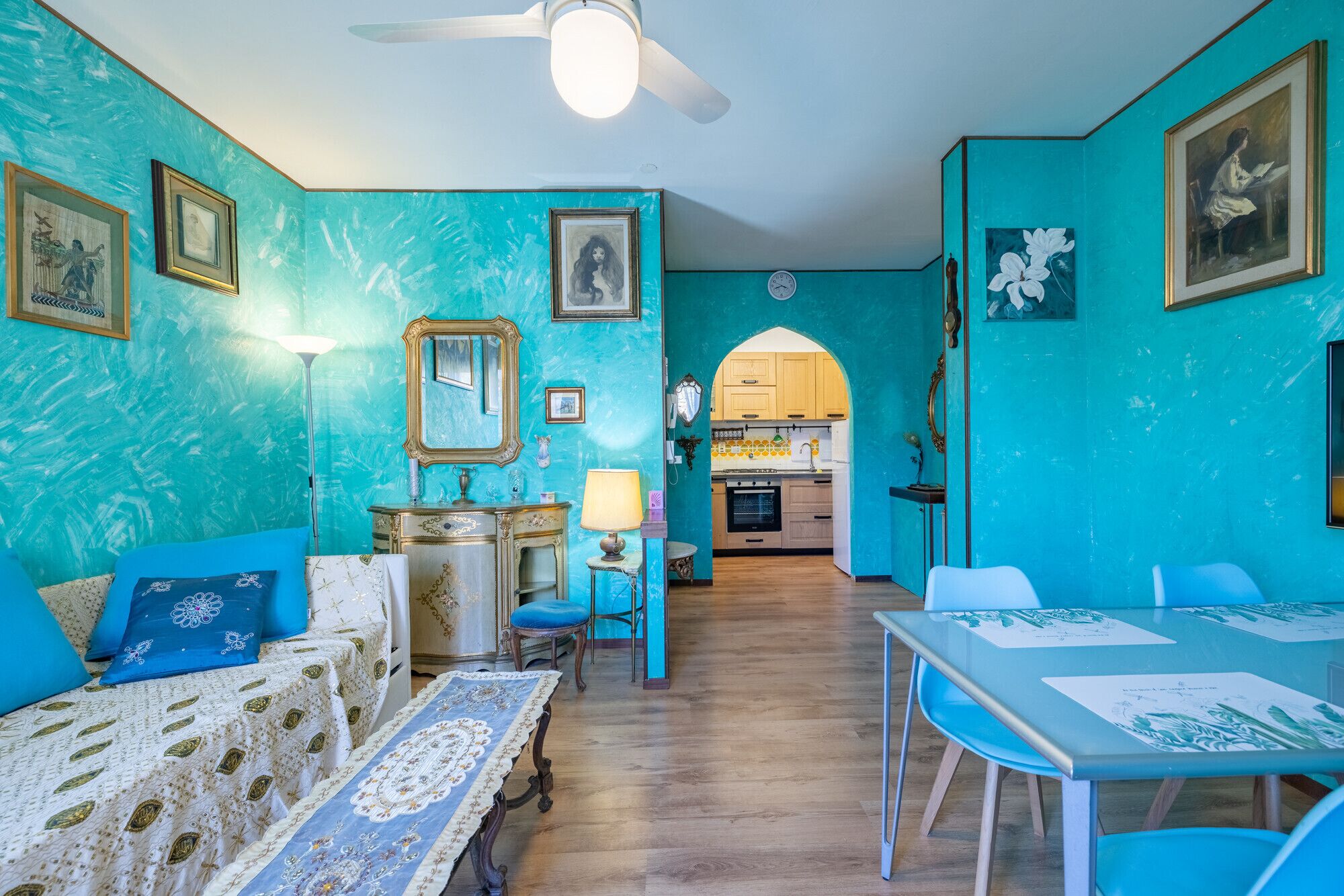 Ein Zimmer mit einer türkisblauen Wand, einem Essbereich mit Tisch und Stühlen, einem Sofa mit gemusterten Kissen und einer durch einen Bogen sichtbaren Küche.