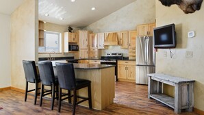 Una cocina con armarios de madera, una isla central con sillas, un televisor montado en la pared y una cabeza de animal decorativa.