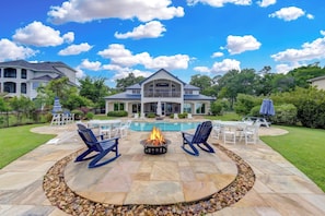 A poolside area with a fire pit, seating, and a large house in the background. - Villa de Lago! A Stunning Waterfront Estate on Lake Conroe (Montgomery)