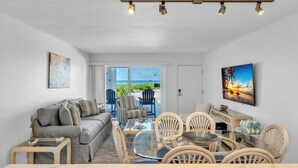 Una habitación con sofá, sillas, una mesa y televisor. Hay una puerta que da a un balcón con vista al océano.