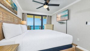 Ein Hotelzimmer mit Bett, Deckenventilator, Fernseher, Fenster mit Meerblick und einem hölzernen Kopfteil.