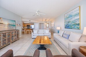 A spacious living room with a wooden coffee table, a sofa, and a dining area with a kitchen in the background. - Phoenix X 407 3 Bedroom Condo (Orange Beach)