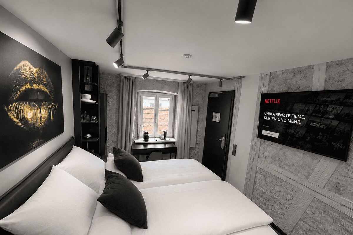 Habitación de hotel con una cama, un escritorio, una estantería, una pantalla de televisión y una ventana.