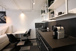 Una cocina moderna con encimera negra, armarios blancos, una cafetera y una zona de comedor con mesa blanca y sillas negras.