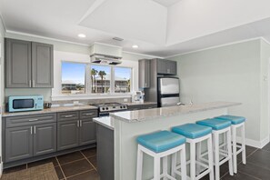 A modern kitchen with grey cabinets, a stainless steel refrigerator, a microwave, a stove, and a large window with a view of palm trees.