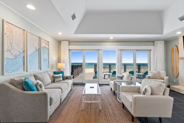 A spacious living room with a large glass door leading to a balcony with a view of the beach. There are two sofas, a coffee table, and a TV.