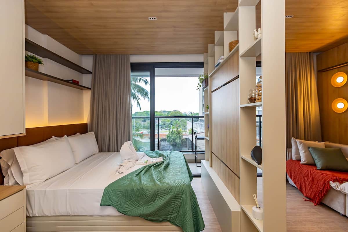 Una habitación de hotel moderna con cama, sofá, un amplio ventanal y estanterías empotradas.