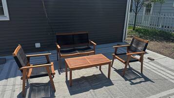 A patio with a wooden table and chairs, a bench, and a house with a dark siding exterior.