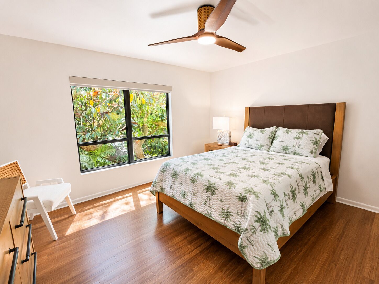 A bedroom with a bed, a nightstand, a lamp, a ceiling fan, a window with a view of trees, and a chair.