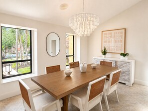 A dining room with a wooden table, white chairs, a chandelier, a mirror, and a view of trees outside. - Renovated 3/3.5 Pool home, Private Beach Access (Longboat Key)