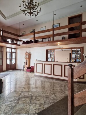 Un hall d’entrée spacieux avec un plafond haut, un grand lustre, un imposant escalier en bois et une horloge traditionnelle.
