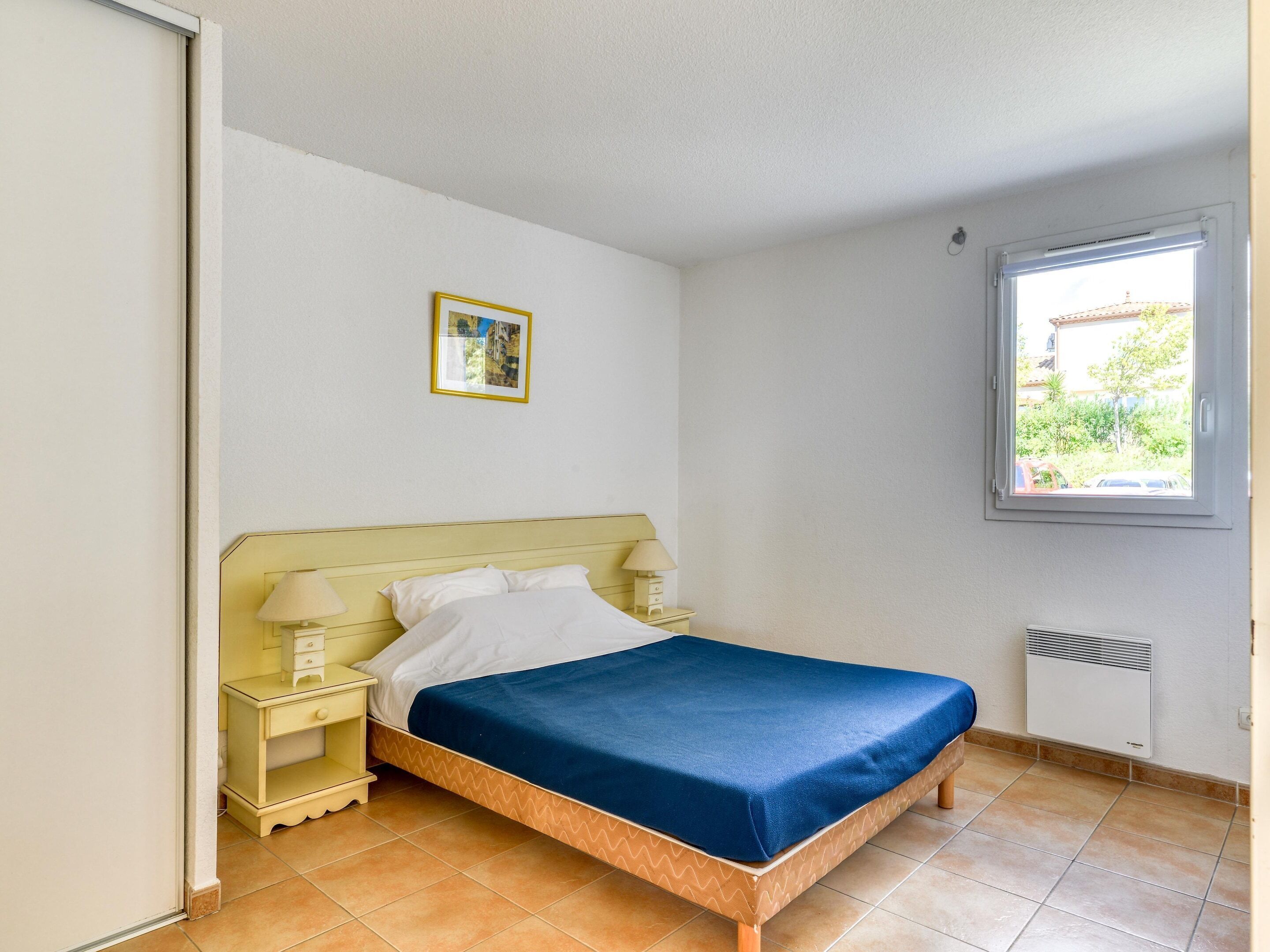 Ein Einzelbett mit blauer Bettdecke, ein Nachttisch mit Lampe, ein Bild an der Wand und ein Fenster mit Blick auf Grünfläche.