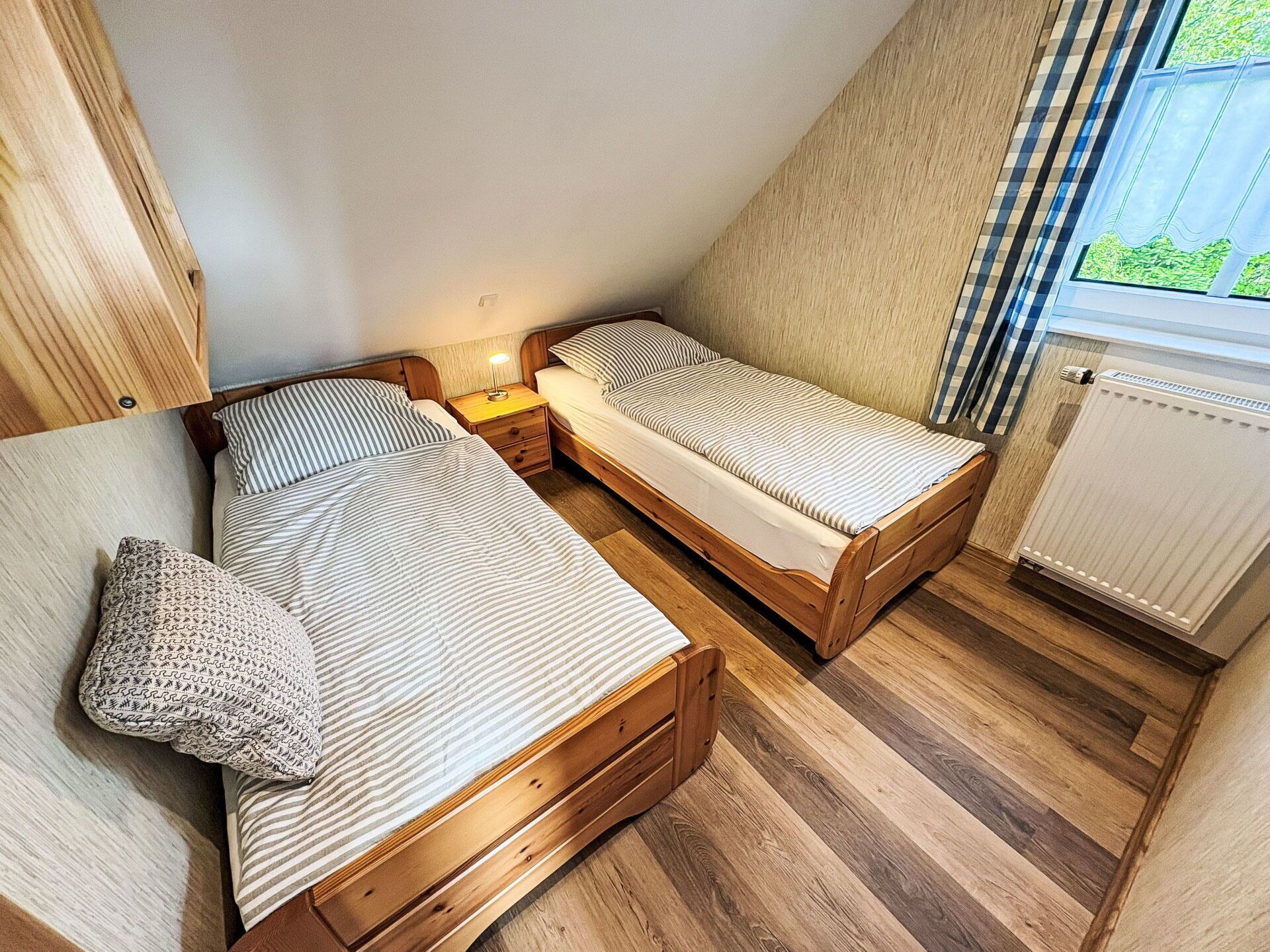 Una habitación pequeña y acogedora con dos camas de madera, una mesita de noche con lámpara y una ventana con vistas a zonas verdes.