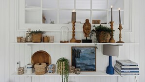 Ein Zimmer mit weißen Wänden, einem großen Fenster, Regalen mit Dekoration und einer Holztür, sowie einem Gemälde.